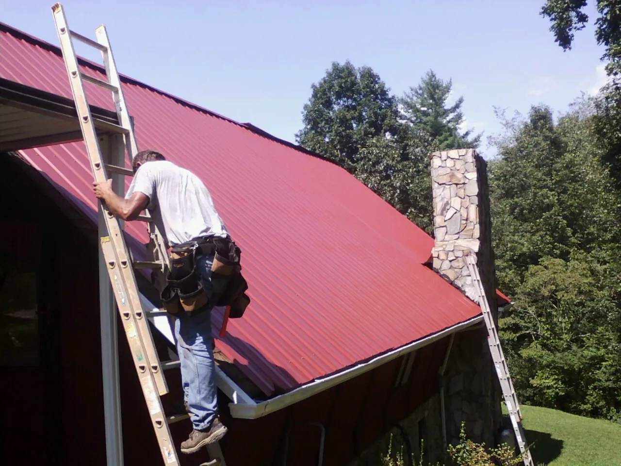Roofer on a ladder beside a red metal roof and stone chimney — Soffit & Fascia Repair by Roofing Brothers in Alachua